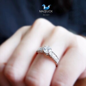 May include: A silver ring with a large, clear, round diamond in the center. The ring is set with smaller diamonds around the center stone and on the band. The ring is on a person's finger.