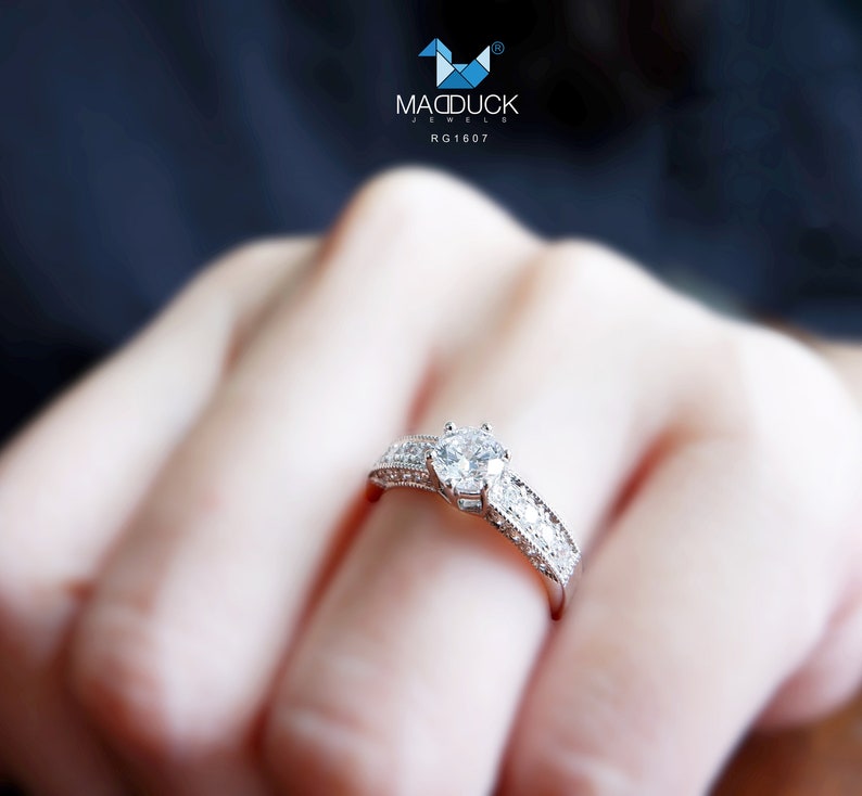 May include: A silver ring with a large, clear, round diamond in the center. The ring is set with smaller diamonds around the center stone and on the band. The ring is on a person's finger.