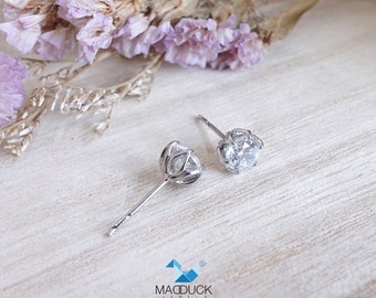 May include: A pair of silver stud earrings with a clear, round gemstone set in a delicate, prong setting. The earrings are on a white wooden surface with dried flowers in the background.
