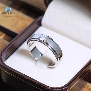 May include: A silver band with a single, round diamond set in the center. The band is polished and has a slightly raised edge. The ring is in a white box with a brown exterior.