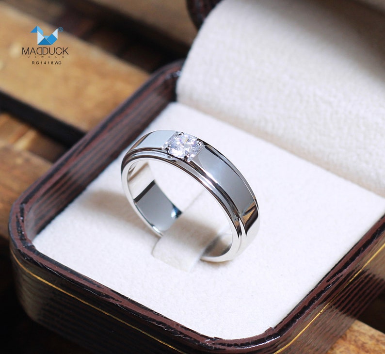 May include: A silver band with a single, round diamond set in the center. The band is polished and has a slightly raised edge. The ring is in a white box with a brown exterior.