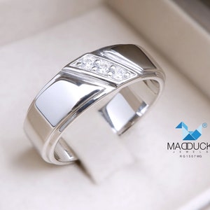 May include: A silver band with a row of small diamonds set in a channel setting. The band has a bevelled edge and a polished finish. The ring is in a white box with the text "MADDUCK JEWELS RG1507WG".