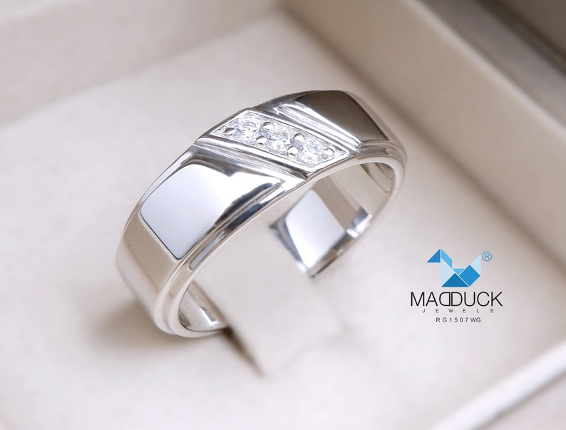 May include: A silver band with a row of small diamonds set in a channel setting. The band has a bevelled edge and a polished finish. The ring is in a white box with the text "MADDUCK JEWELS RG1507WG".