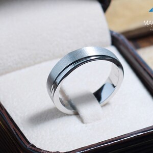 May include: A silver ring with a brushed finish and a polished inner band, presented in a brown jewelry box with a white interior. The ring is from MacDuck Jewels, model RG1728WGR.