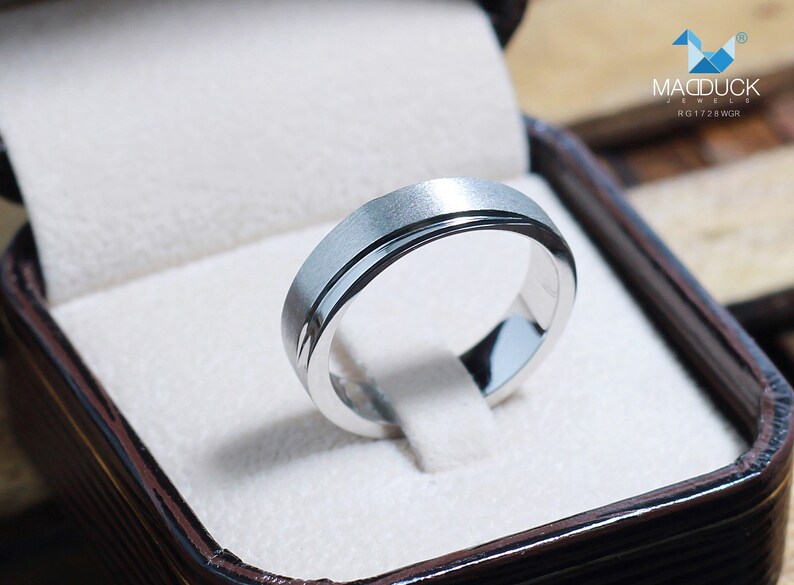 May include: A silver ring with a brushed finish and a polished inner band, presented in a brown jewelry box with a white interior. The ring is from MacDuck Jewels, model RG1728WGR.
