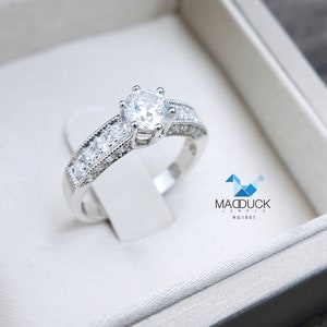 May include: A silver engagement ring with a large round center stone and smaller stones set in a band. The ring is in a white box with the text "Madduck Jewels RG1607".