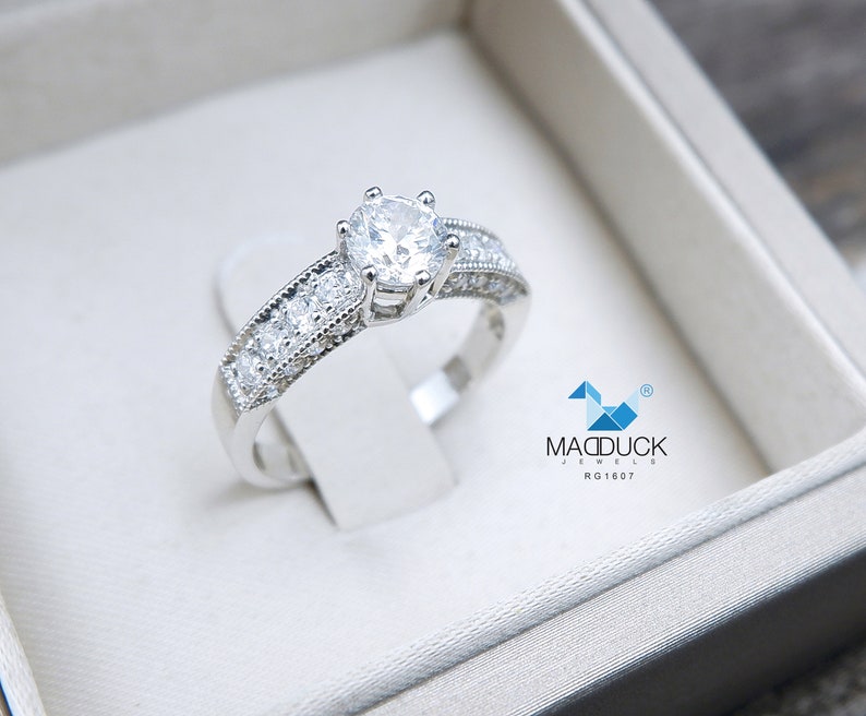 May include: A silver engagement ring with a large round center stone and smaller stones set in a band. The ring is in a white box with the text "Madduck Jewels RG1607".