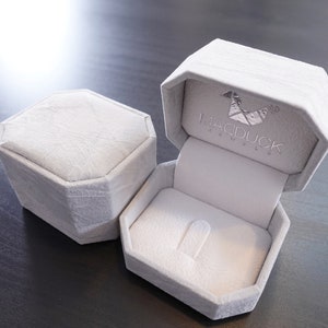 May include: A white ring box with a gray velvet interior. The box is open and the lid has the text "MADDUCK JEWELLERY" printed on it.