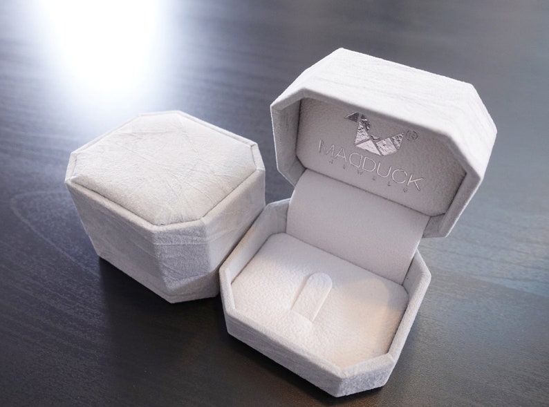 May include: A white ring box with a gray velvet interior. The box is open and the lid has the text "MADDUCK JEWELLERY" printed on it.