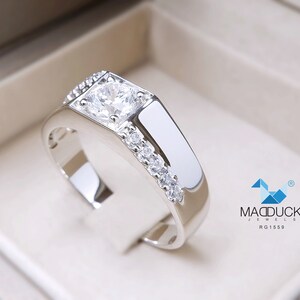 May include: A silver ring with a large, clear, square-cut gemstone surrounded by smaller gemstones. The ring is set in a box with the logo "MADDUCK JEWELS RG1559".