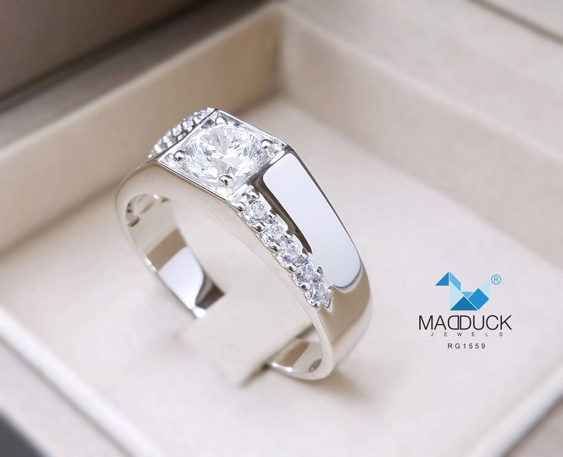 May include: A silver ring with a large, clear, square-cut gemstone surrounded by smaller gemstones. The ring is set in a box with the logo "MADDUCK JEWELS RG1559".