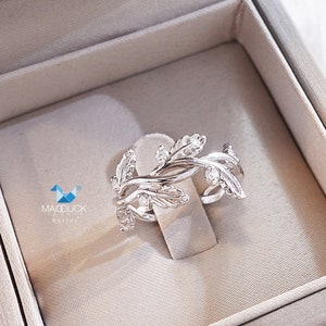 May include: A silver ring with a delicate design of leaves and small clear stones. The ring is in a white velvet ring box.