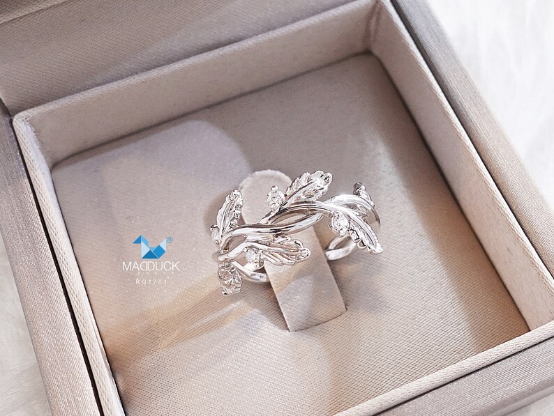 May include: A silver ring with a delicate design of leaves and small clear stones. The ring is in a white velvet ring box.