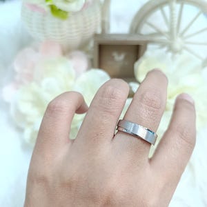 May include: A silver ring with a brushed finish is worn on a finger. The ring has a flat, wide band with a subtle, textured surface. The background includes flowers and decorative elements.