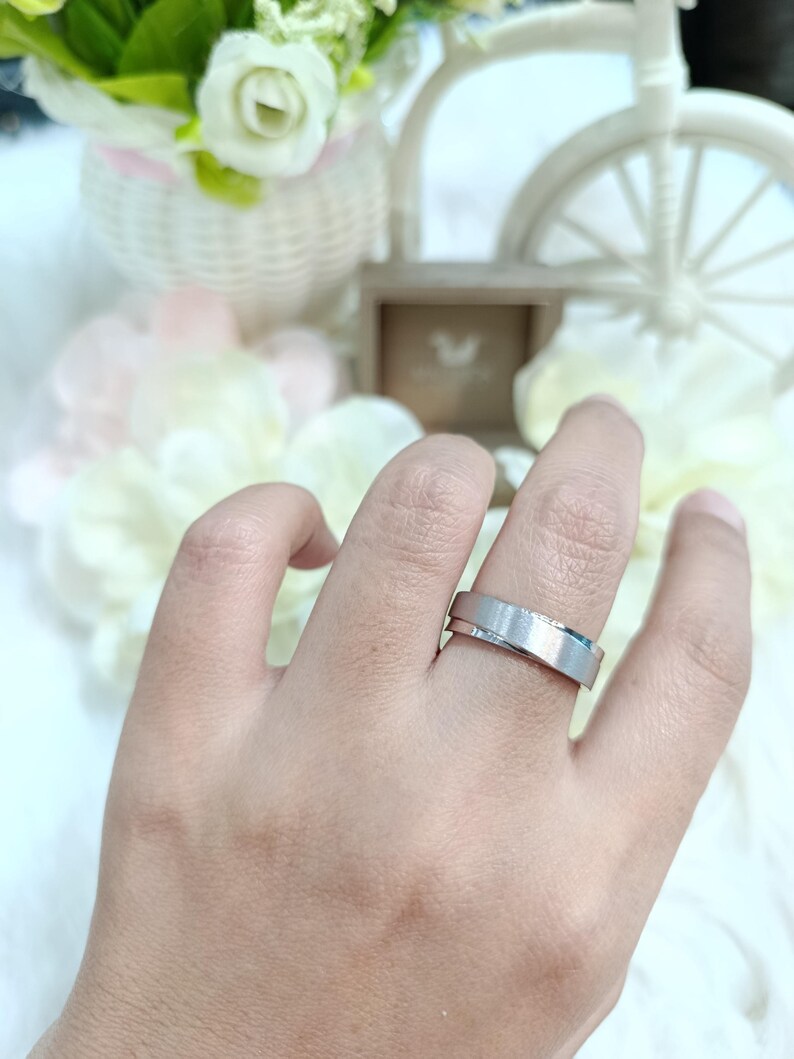 May include: A silver ring with a brushed finish is worn on a finger. The ring has a flat, wide band with a subtle, textured surface. The background includes flowers and decorative elements.