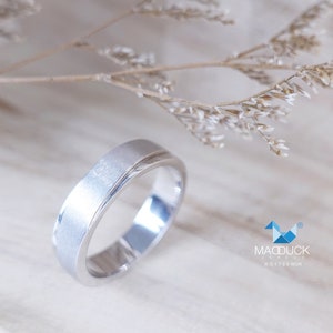 May include: A silver ring with a brushed finish and a polished inner band. The ring is set against a soft, neutral background with dried floral accents. The ring is a simple, elegant design.