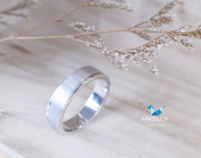 May include: A silver ring with a brushed finish and a polished inner band. The ring is set against a soft, neutral background with dried floral accents. The ring is a simple, elegant design.