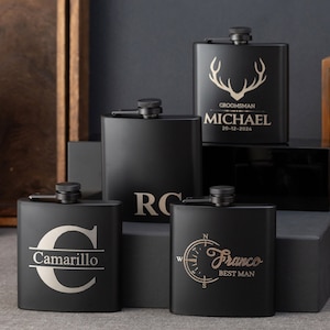 Könnte beinhalten: Vier schwarze Metall-Hüftflaschen mit gravierten Designs. Die Flaschen haben einen Klappverschluss und sind mit Namen und Text personalisiert. Die Designs beinhalten einen Kompass, einen großen Buchstaben C, einen Hirsch und den Text "RG".