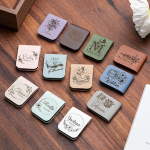 May include: A collection of 11 personalised leather bookmarks with various designs and names. The bookmarks are in different colours, including brown, blue, green, and pink. Each bookmark features a unique design, such as flowers, books, and a jar with flowers. The bookmarks are a great way to personalise your reading experience.