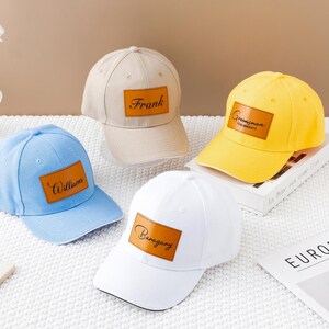 以下が含まれることがあります： ライトブルー、ベージュ、イエロー、ホワイトの野球帽4つ。各キャップには、名前が印刷された茶色の長方形のパッチが付いています。キャップは、ニュートラルな背景の白い表面に表示されています。