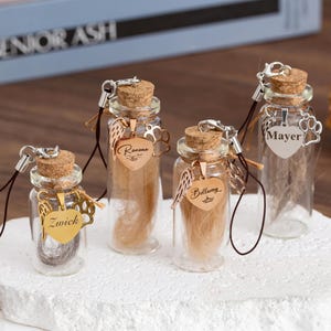 May include: Four small glass memorial bottles with cork stoppers, each containing hair or ash. The bottles have decorative charms, including angel wings and heart-shaped pendants with names. The bottles sit on a white surface.