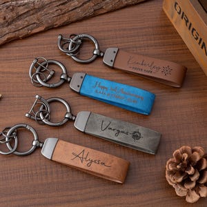 May include: Four personalised keychains with metal rings and leather straps. The straps are brown, blue, and grey, each engraved with names and messages. One keychain reads "Kimberley - DRIVE SAFE", another says "Happy 5th Anniversary", and the others have names.
