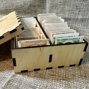 May include: A wooden tea bag storage box with a lid. The box is filled with tea bags and has a black trim. The lid has the word "Tea" engraved on it.