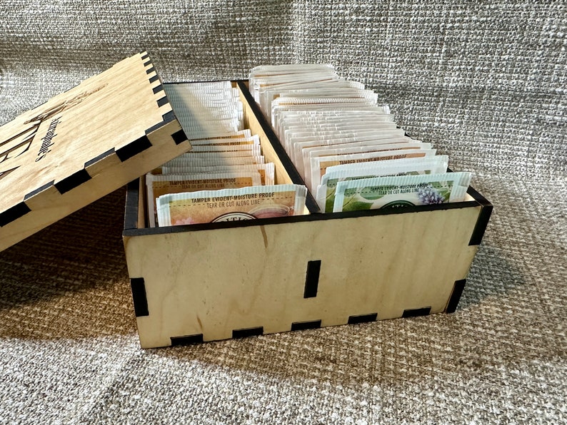 May include: A wooden tea bag storage box with a lid. The box is filled with tea bags and has a black trim. The lid has the word "Tea" engraved on it.