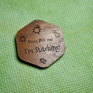 May include: A brown, wooden, hexagonal craft accessory with the text "Don't BUG me! I'm Stitching!" and bug illustrations. It is set against a green, textured background.