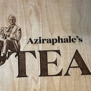 May include: A wood sign with a black and white image of a man holding a teacup. The sign says "Aziraphale's TEA" in black lettering.