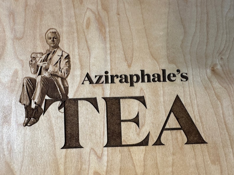 May include: A wood sign with a black and white image of a man holding a teacup. The sign says "Aziraphale's TEA" in black lettering.