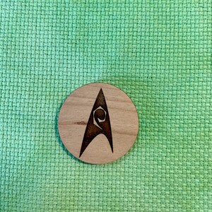May include: A round wooden coaster with a laser-etched design. The design features a stylized letter "A" with a unique symbol inside. The coaster is made of light-colored wood and sits on a green textured surface.