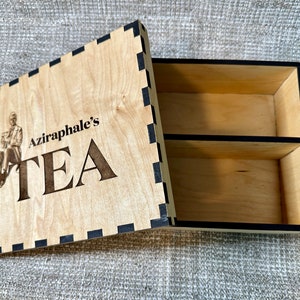 May include: A wooden tea box with a black and white checkered border. The lid features a laser-engraved image of a man in a coat and the text "Aziraphale's TEA".