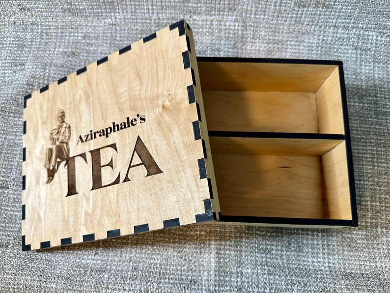 May include: A wooden tea box with a black and white checkered border. The lid features a laser-engraved image of a man in a coat and the text "Aziraphale's TEA".
