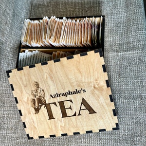 May include: A wooden box with a black trim and a black and white illustration of a person. The box has the text "Aziraphale's TEA" on the front.