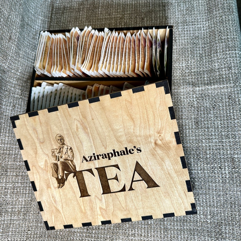 May include: A wooden box with a black trim and a black and white illustration of a person. The box has the text "Aziraphale's TEA" on the front.
