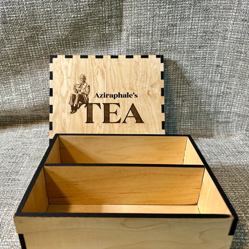 May include: A wooden tea box with a black trim and a lid that features a drawing of a man in a suit holding a cup. The lid also has the text "Aziraphale's Tea" engraved on it.
