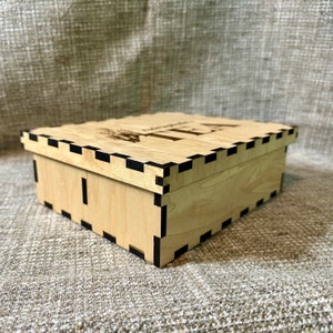 May include: A wooden box with a black trim and the word "TEA" engraved on the top. The box is made of light wood and has a simple design.