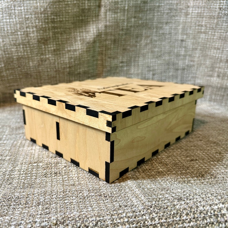 May include: A wooden box with a black trim and the word "TEA" engraved on the top. The box is made of light wood and has a simple design.