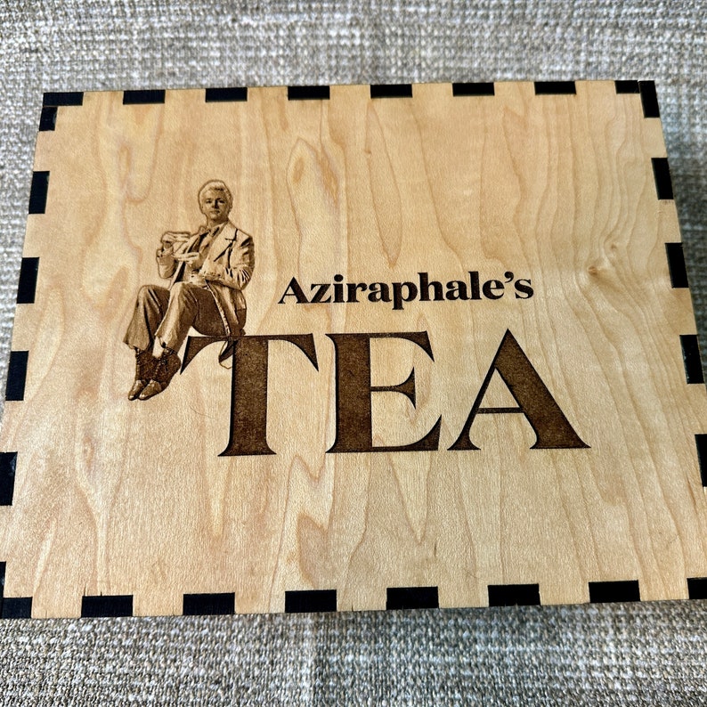 May include: A wooden box with a black border and a laser-engraved image of a man holding a teacup. The text "Aziraphale's TEA" is also engraved on the box.
