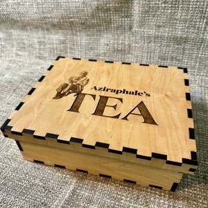 May include: A wooden tea box with a black border. The box has an image of a man sitting and the text "Aziraphale's TEA" is engraved on the lid.