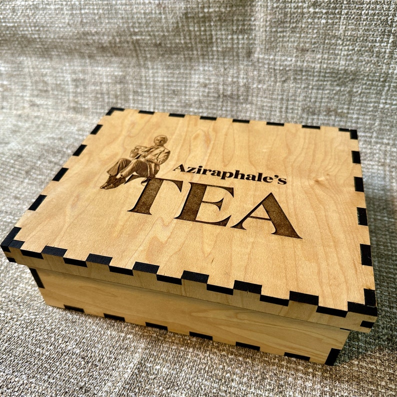 May include: A wooden tea box with a black border. The box has an image of a man sitting and the text "Aziraphale's TEA" is engraved on the lid.