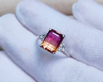 May include: A silver ring with a large, rectangular, multi-colored gemstone. The gemstone is a gradient of pink, orange, and yellow. The ring is set with small, clear gemstones on the sides.