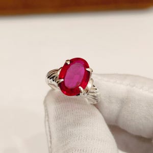 May include: A silver ring featuring a large, oval-cut ruby gemstone. The ruby is a vibrant red color, and the ring has a textured band. The ring is held by a gloved hand.