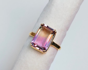 May include: A gold ring with a large, rectangular, iridescent gemstone. The gemstone is a mix of pink, purple, and yellow.