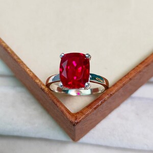May include: A silver ring featuring a large, cushion-cut, vibrant red gemstone. The gemstone is the focal point, set on a simple, polished silver band. The ring is displayed in a brown wooden box.