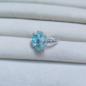 May include: A silver ring with a blue topaz flower design. The ring has a delicate band and a cluster of blue topaz stones set in a flower shape.