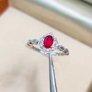 Op de afbeelding: Een zilveren ring met een rode ovale edelsteen omringd door kleinere witte edelstenen. De ring heeft een delicaat, ingewikkeld ontwerp.