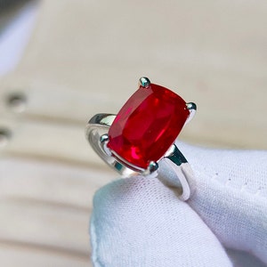 May include: A silver ring featuring a large, rectangular, vibrant red gemstone. The gemstone is held in place by four prongs. The ring is set against a soft, white background, highlighting the brilliance of the stone.
