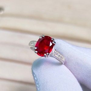 Può includere: Un anello d'argento con una grande gemma rossa brillante a taglio ovale. La fascia dell'anello è decorata con piccole pietre di accento trasparenti. L'incastonatura ha un design classico ed elegante, che mette in risalto il rubino centrale.